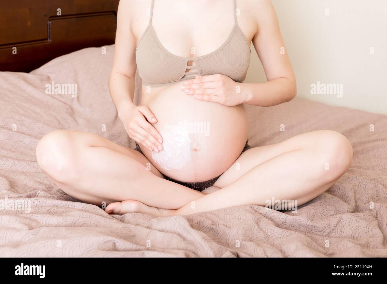 una niña embarazada se sienta en casa en la cama y le mancha una crema antiestrías en el estómago. El embarazo, la maternidad, la preparación y la expectativa conce Foto de stock