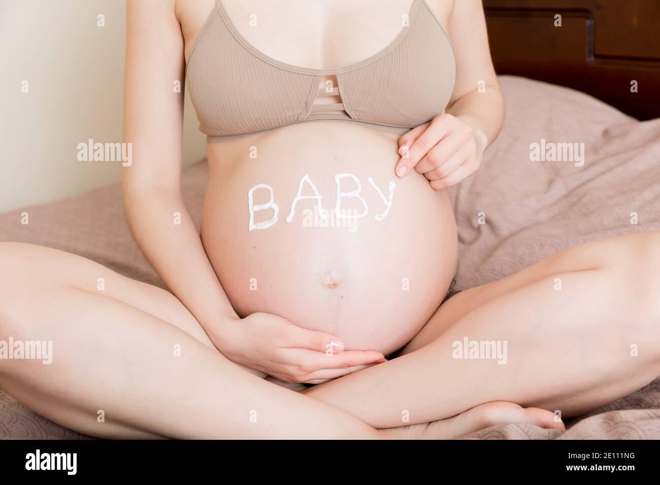una niña embarazada se sienta en casa en la cama y le mancha una crema antiestrías en el estómago. El embarazo, la maternidad, la preparación y la expectativa conce Foto de stock