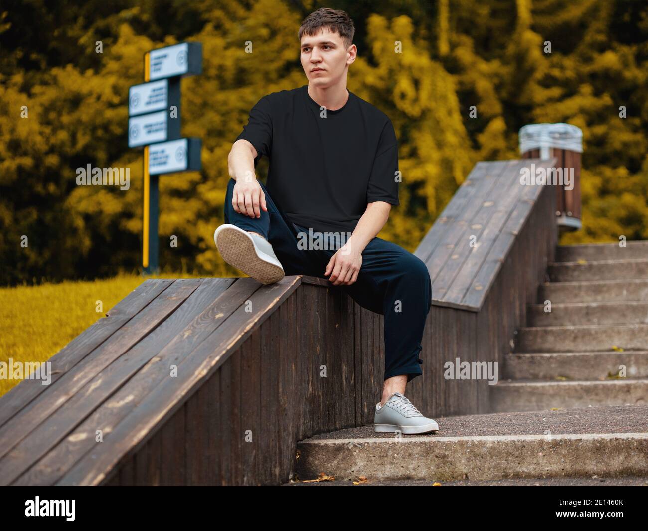 Burlándose de una camiseta negra en un chico con pantalones y zapatillas de  deporte sentado en un parque sobre un fondo borroso de árboles, vista  frontal. Ropa de Marca informal Fotografía de