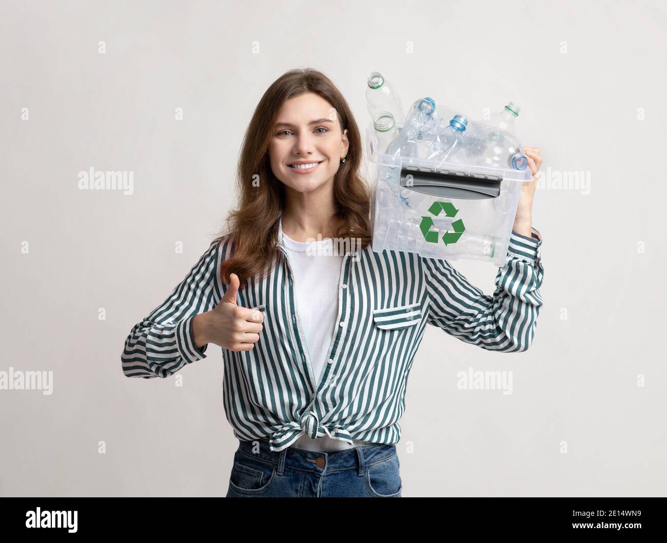 Mujer sonriente sosteniendo papelera de reciclaje con botellas de plástico y mostrando pulgar hacia arriba Foto de stock