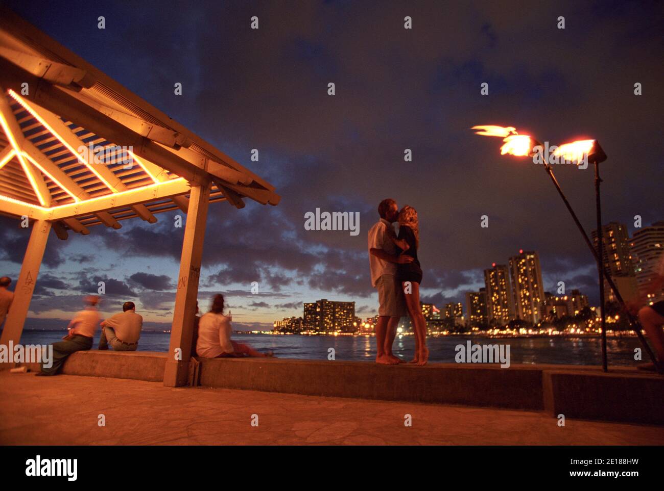 Pareja besando en un muelle en Waikiki al atardecer con antorchas tiki y luces de hotel en el fondo Foto de stock
