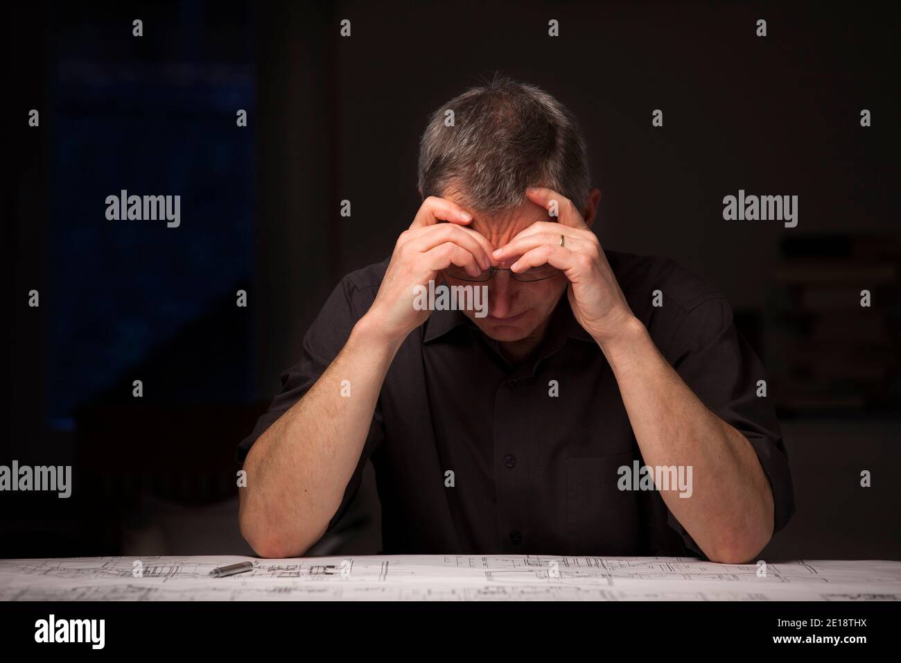 Cansado y sobrertrabajado arquitecto o ingeniero sentado en casa en un escritorio con plano - imagen oscura Foto de stock