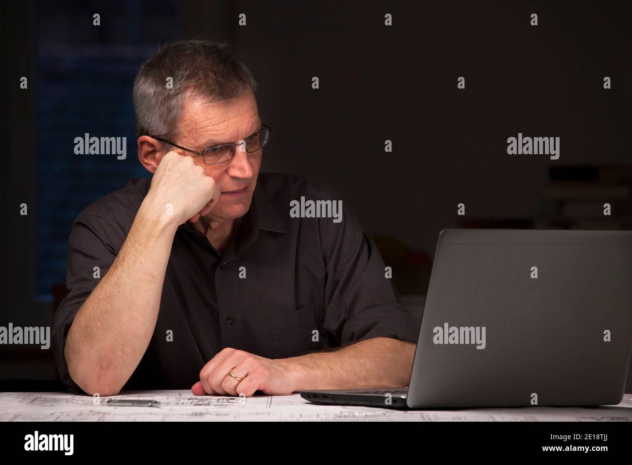 Hombre de negocios maduro o ingeniero en camisa oscura sentado por la noche en un escritorio mirando a un portátil - imagen oscura Foto de stock