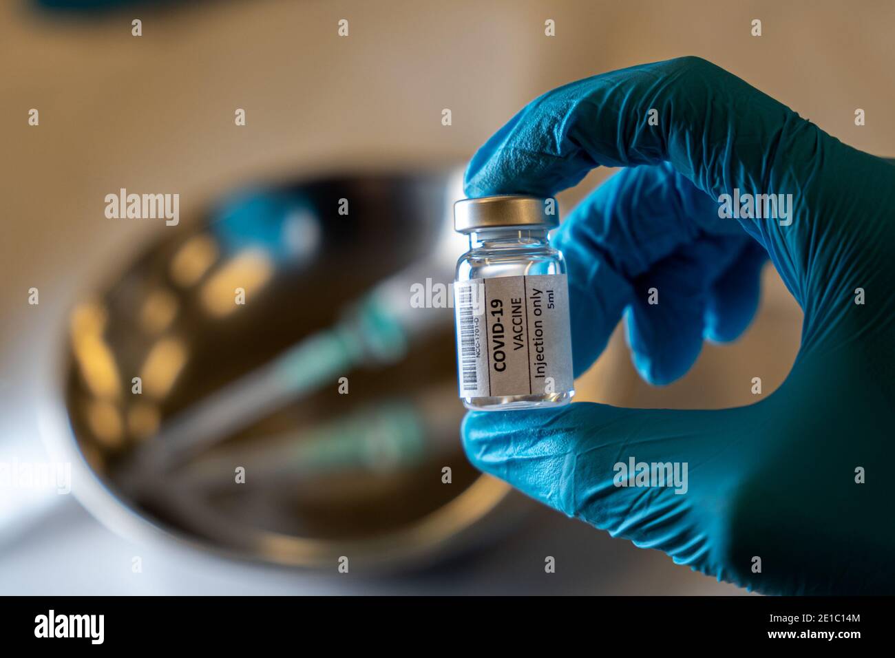 NIJMEGEN, PAÍSES BAJOS - 2 DE ENERO: Vacuna de la corona / ilustración de la vacuna Covid-19. Vial que se sujeta con una mano en guantes azules. Jeringa en una bandeja de metal i Foto de stock