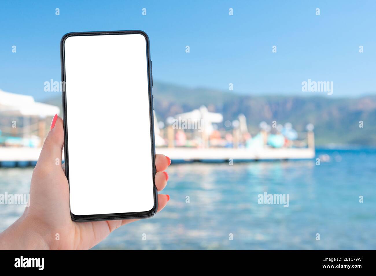Imagen de una mujer que sostiene una mano celular teléfono móvil  inteligente con pantalla de escritorio en blanco junto al mar y el cielo  azul. Concepto de uso de internet para vacaciones