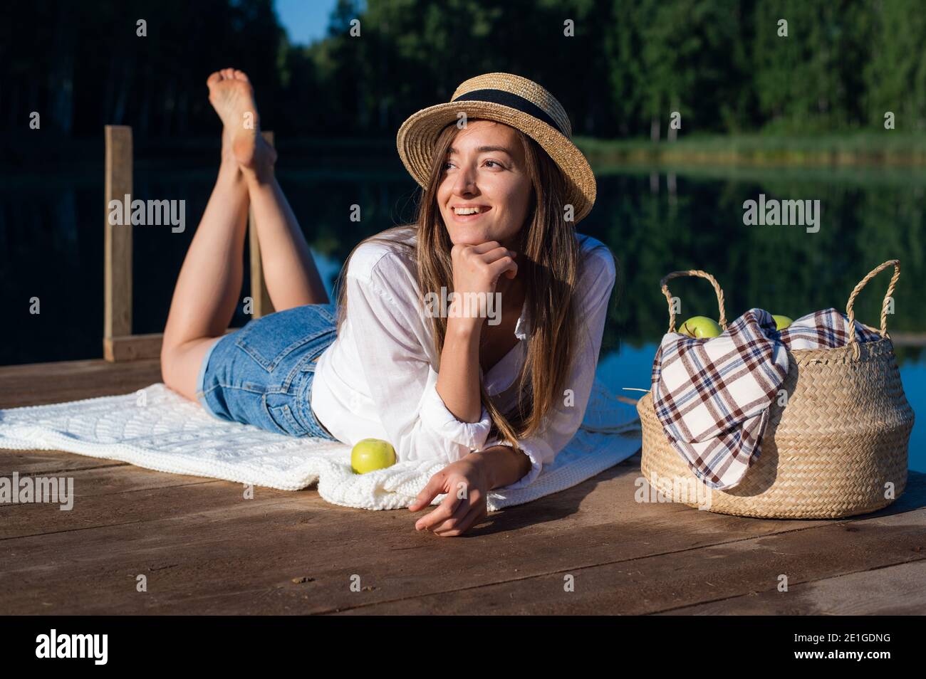 Hermosa mujer joven con pantalones cortos azules, camisa y sombrero  disfruta de su picnic por la mañana en un muelle de madera cerca del lago  Fotografía de stock - Alamy