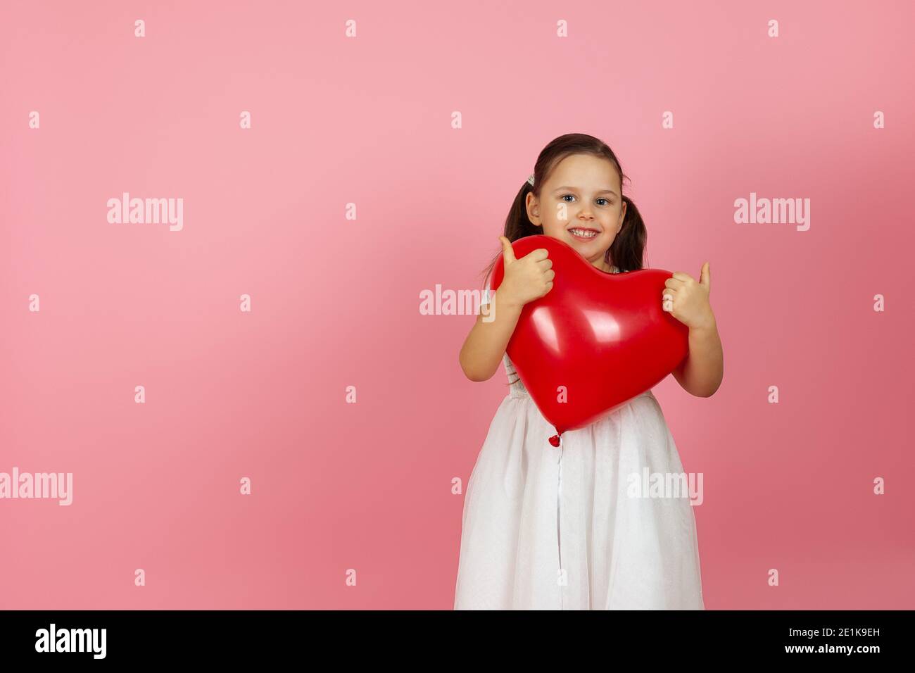 Disfrazarse Perky, chica juguetona con vestido blanco sosteniendo un globo rojo en forma de corazón, dando un pulgares en dos manos, aislado en un fondo rosa Foto de stock