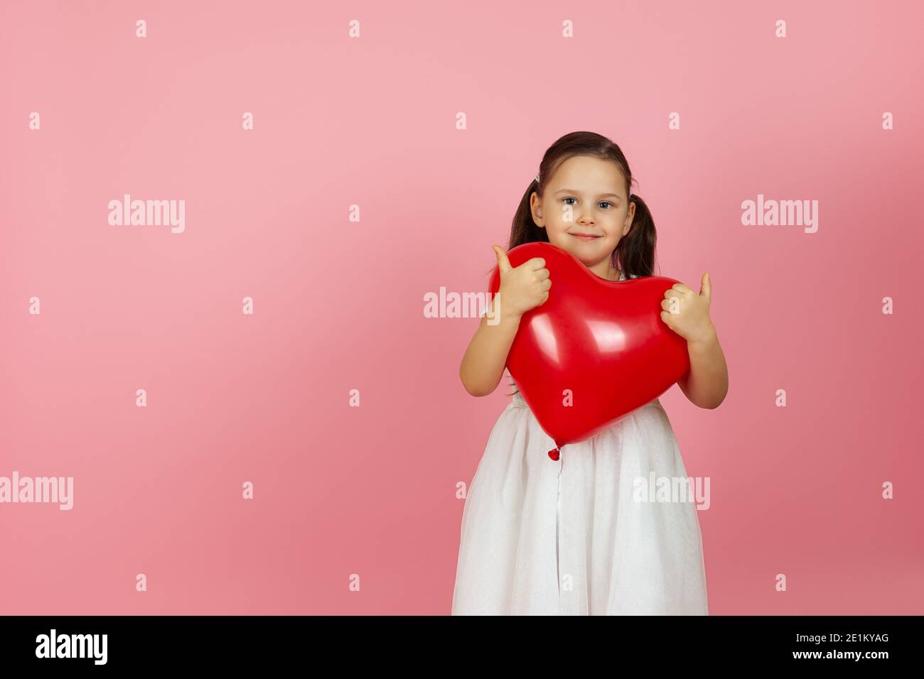 disfrazarse amablemente, niña de corazón en vestido blanco sosteniendo globo rojo en forma de corazón, dando un pulgares en dos manos, aislado en un fondo rosa Foto de stock
