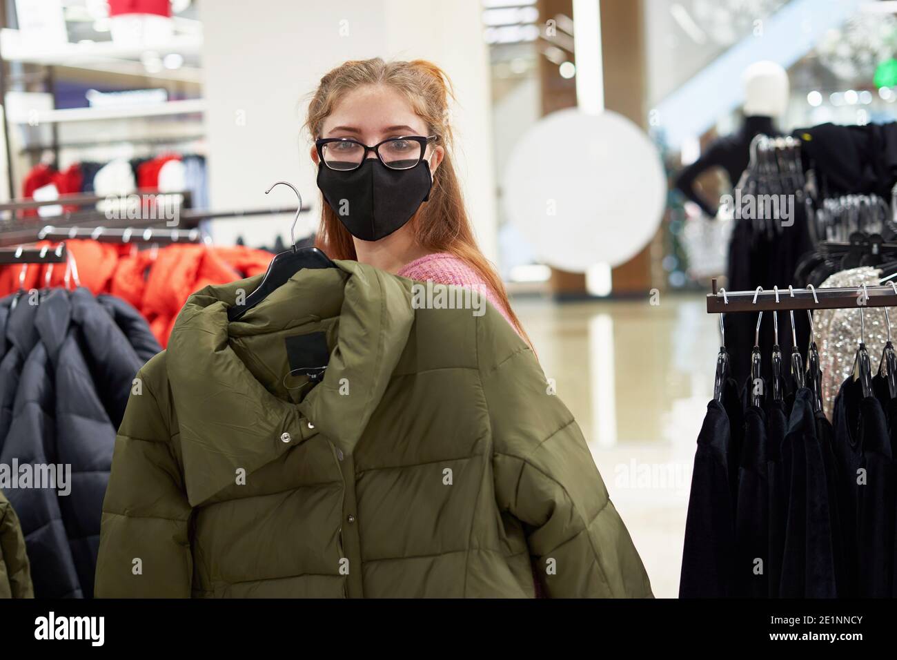 Mujer joven en máscara protectora compra chaqueta de invierno en el almacenar Foto de stock