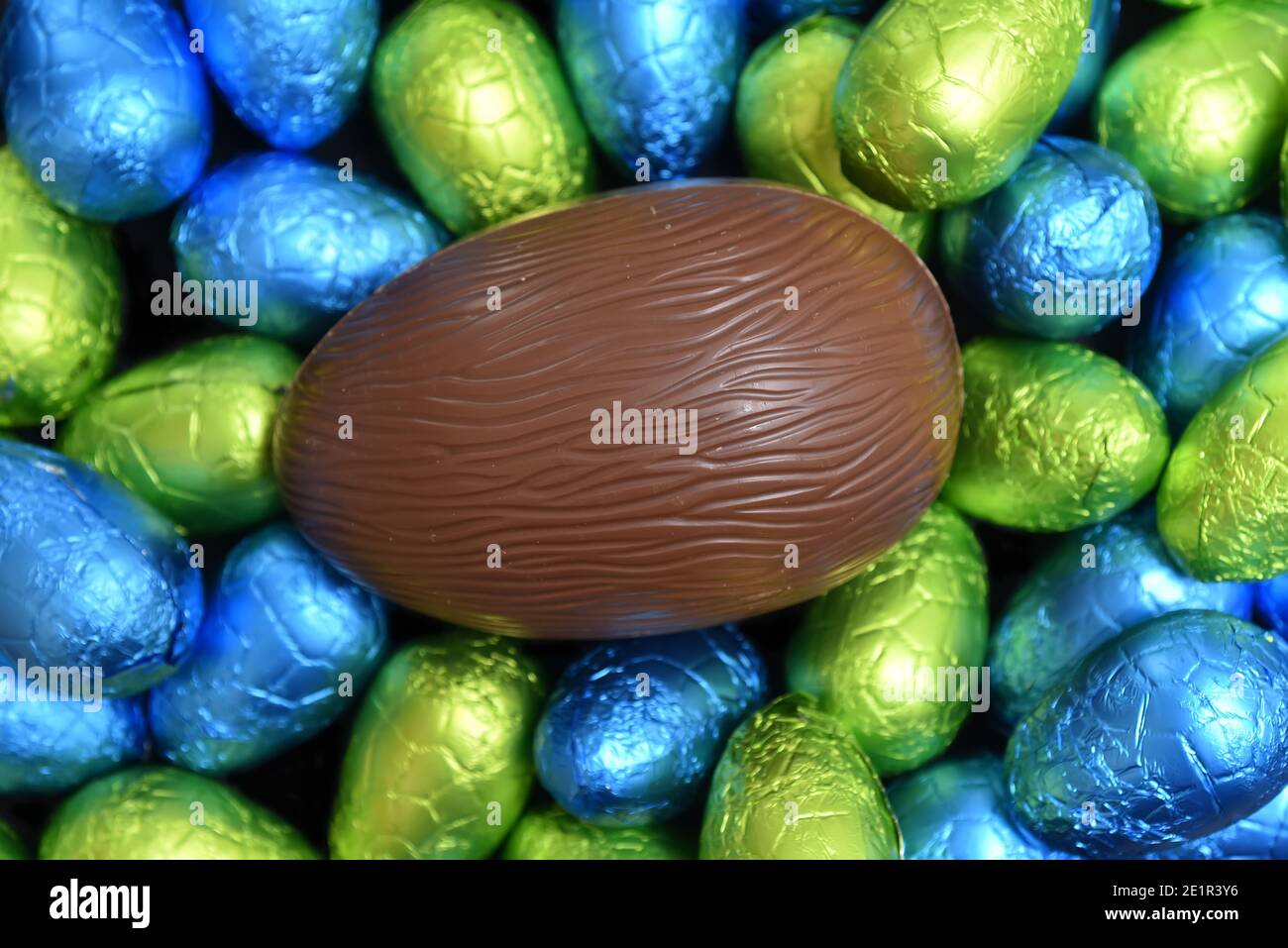 Pila o grupo de varios colores y diferentes tamaños de chocolate envuelto en papel de aluminio de colores huevos de pascua en azul, amarillo y verde lima con una gran m marrón Foto de stock