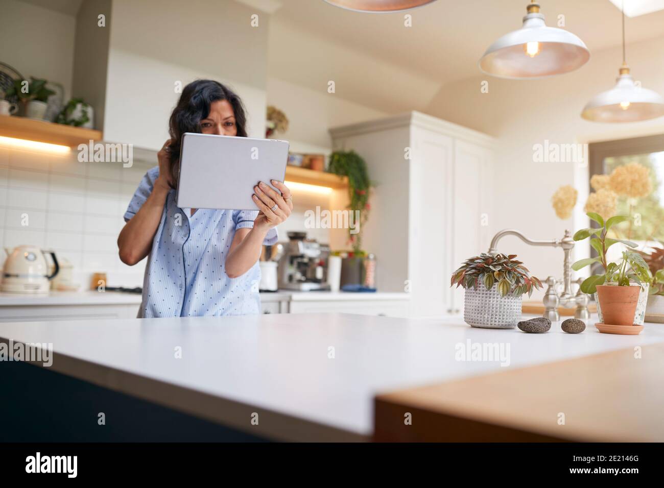 Mujer Hispana madura en pijama en casa en cheques de cocina Cabello y apariencia con tableta digital Foto de stock