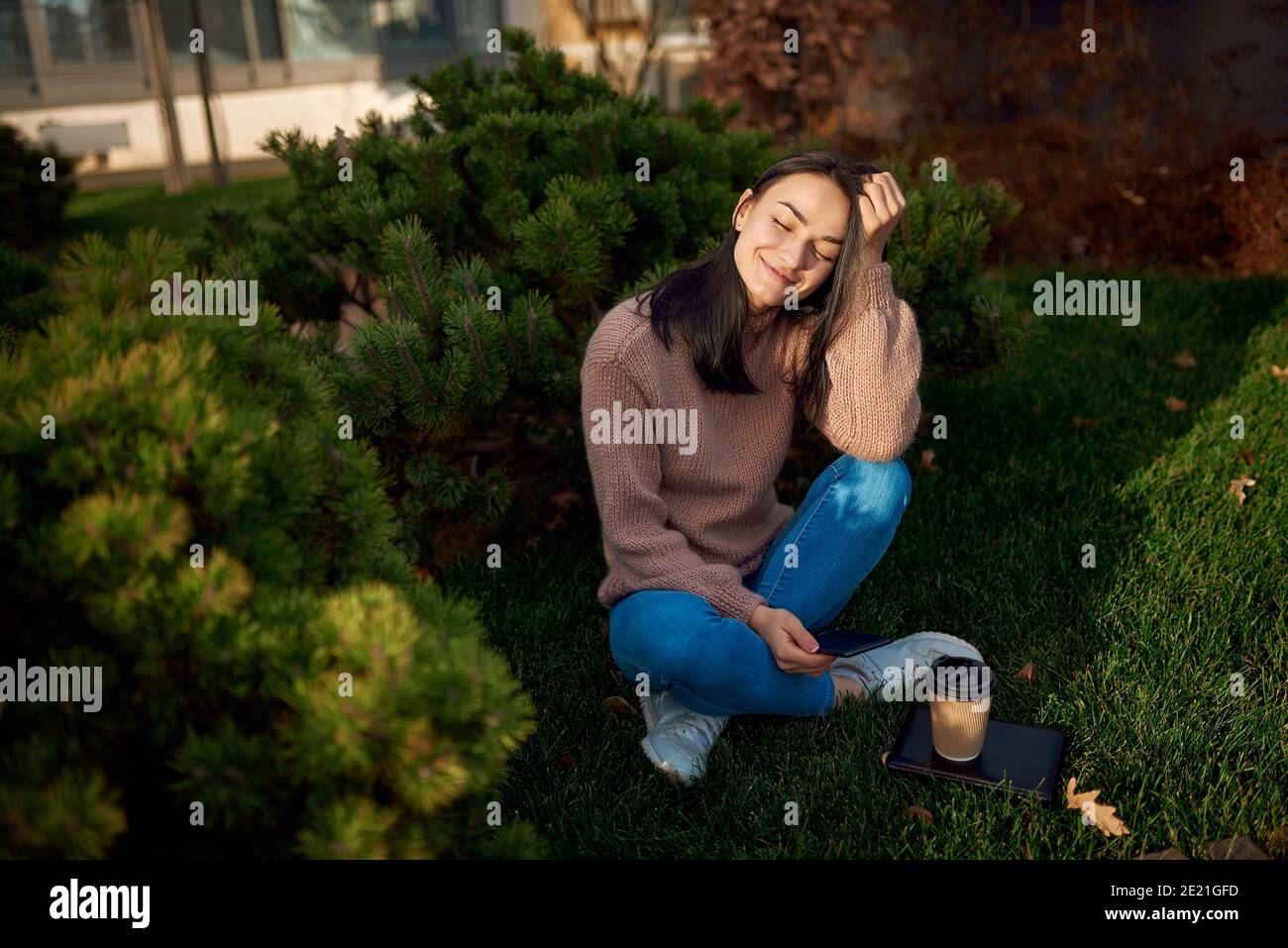 Encantadora señora sentada de piernas cruzadas en el barrio verde y sosteniendo a ella teléfono móvil en la mano Foto de stock