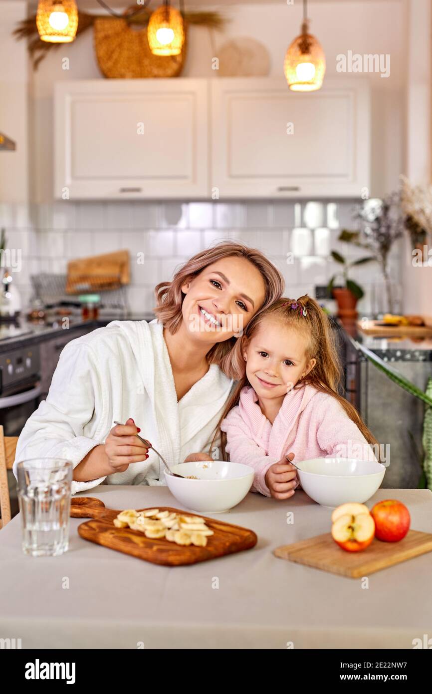 chica disfrutar de la comida para el desayuno con mamá, se sientan juntos la cocina moderna en casa, usando ropa doméstica, comer, miran la cámara smil de stock -