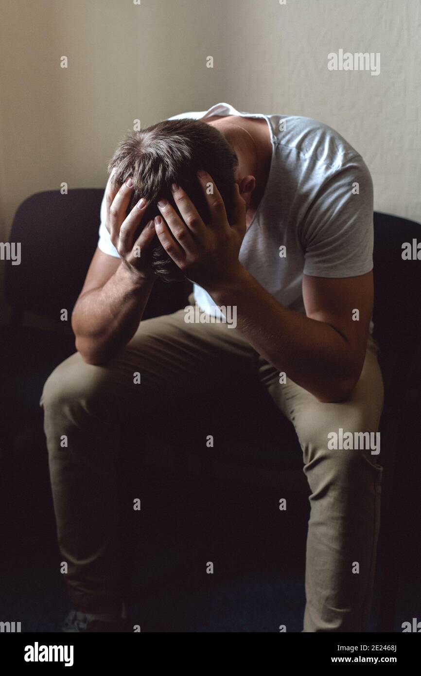 Un joven en estado de crisis y desesperación. El tipo blanco está sentado en una silla y sosteniendo la cabeza con las manos. Foto de stock