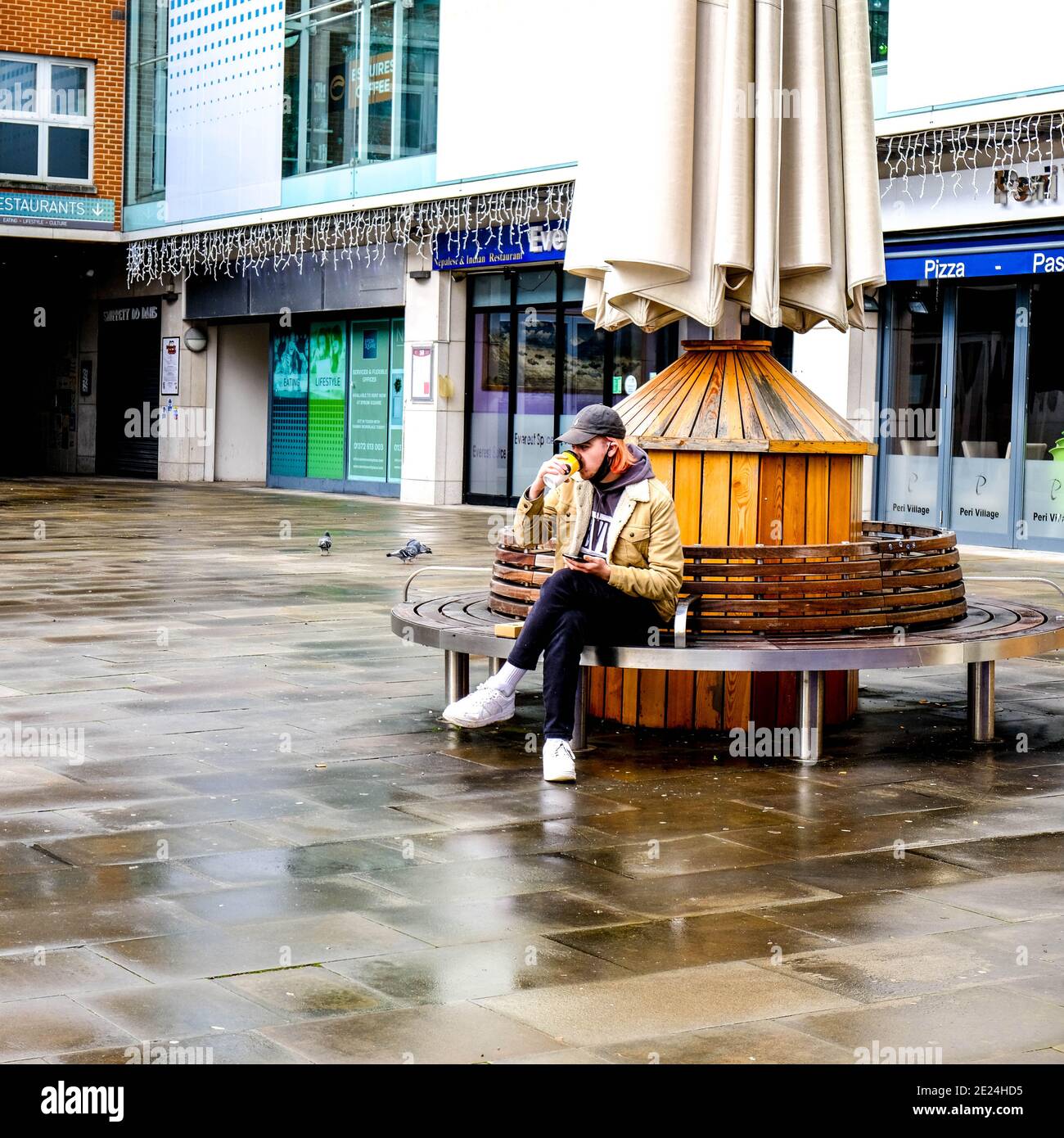 Londres, Reino Unido, 03 2021 de enero, joven sentado solo en un banco de madera redondo tomando café Takeaway durante el cierre pandémico Covid-19 Foto de stock