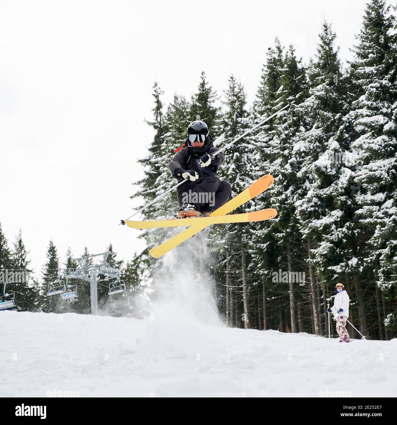 Esquiador masculino en traje de esquí y casco de esquí en nieve fresca en polvo con pinos nevados en el fondo. Hombre freerider en gafas de esquí sosteniendo postes de esquí y haciendo saltar mientras se desliza por pistas cubiertas de nieve Foto de stock