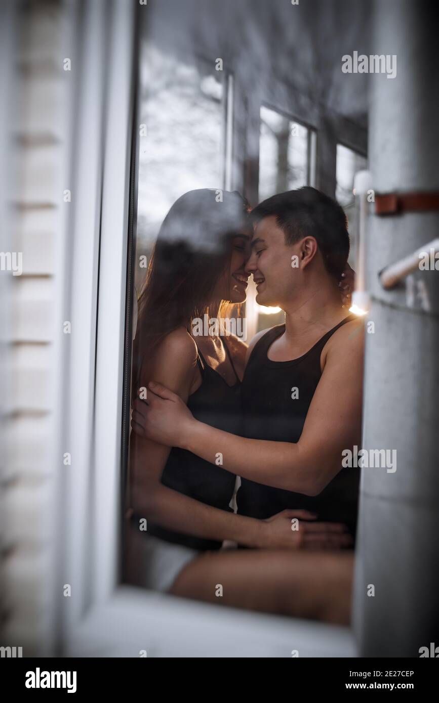 Una pareja joven y cariñosa se abraza en la ventana de su terraza en el apartamento. Un joven se ríe con una hermosa mujer en casa. Los amantes besan, pasan Foto de stock