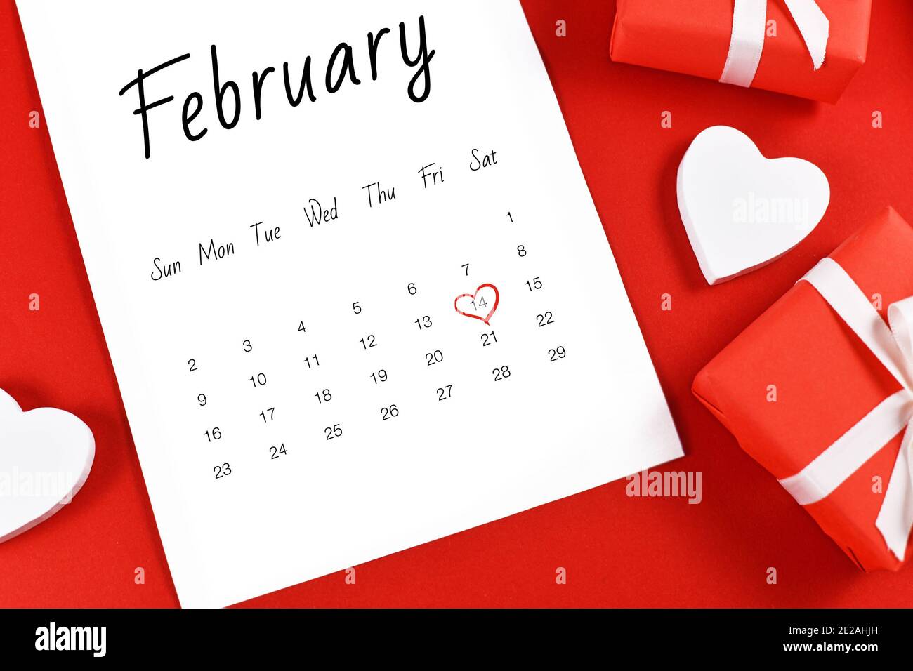 Primer plano de la hoja de calendario de febrero con el día de San Valentín el 14 marcado con corazón rojo rodeado de cajas de regalo y el corazón sobre fondo rojo Foto de stock