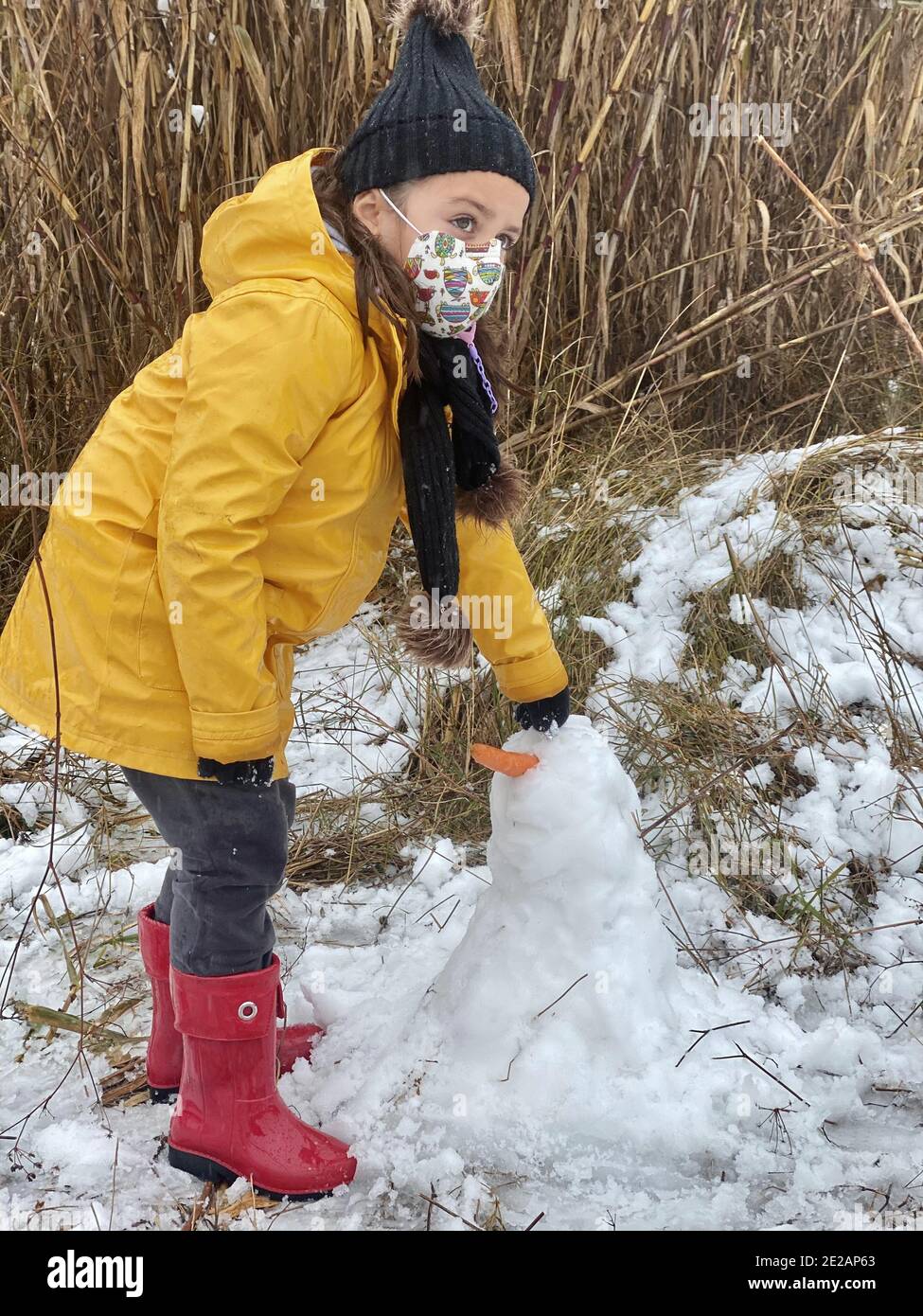 Niña en una máscara protectora y una chaqueta amarilla, poniendo un ojo en  el muñeco de nieve Fotografía de stock - Alamy