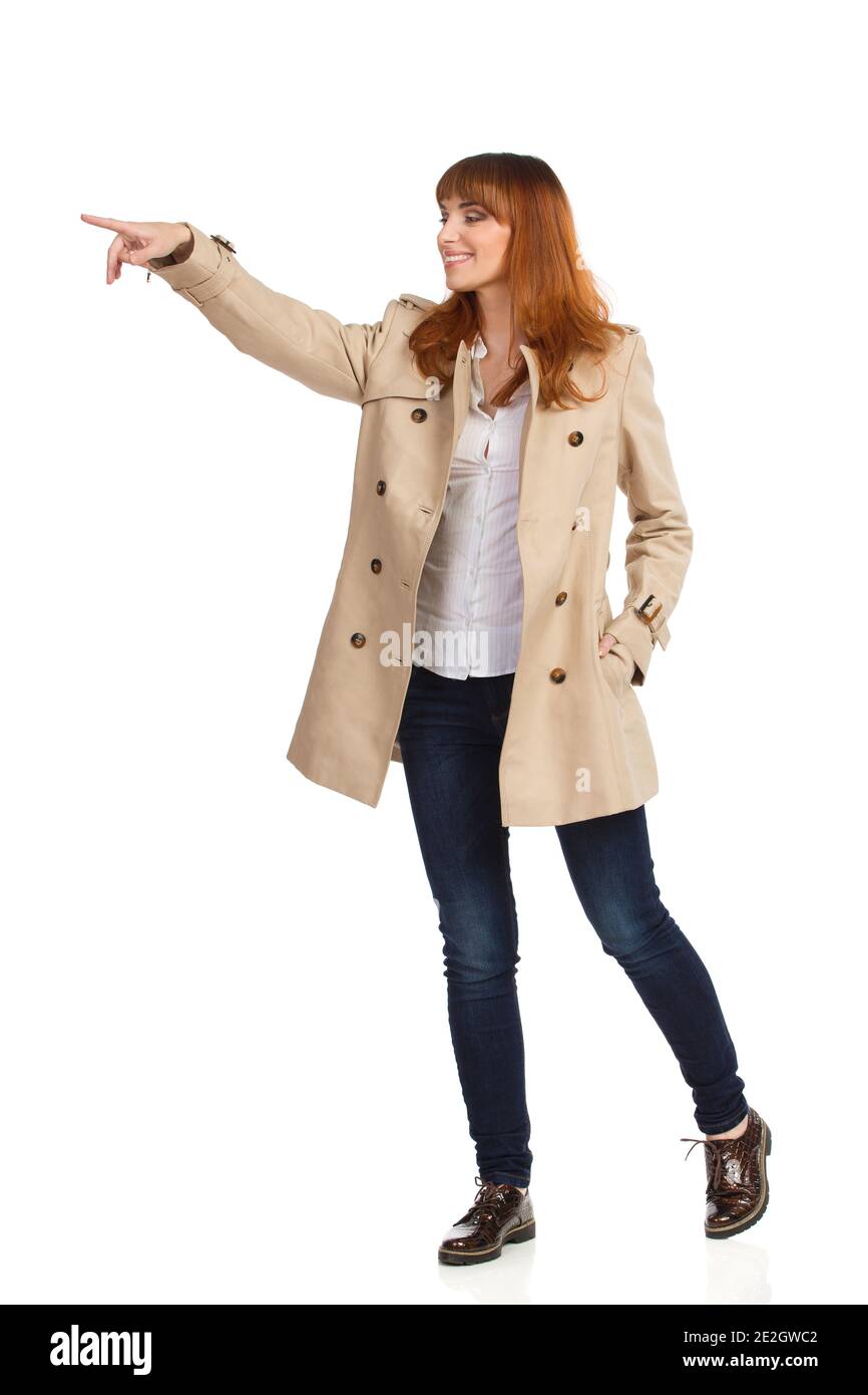 Feliz mujer joven en unbuttoned beige abrigo, jeans y zapatos marrones está  de pie, mirando y señalando. Estudio de duración completa aislado sobre  blanco Fotografía de stock - Alamy
