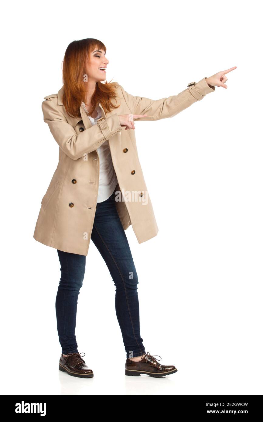 Mujer joven en un abrigo de color beige sin botones, jeans y zapatos marrones está de pie, señalando, mirando y riendo. Estudio de completa, aislado en el muelle Fotografía de stock -