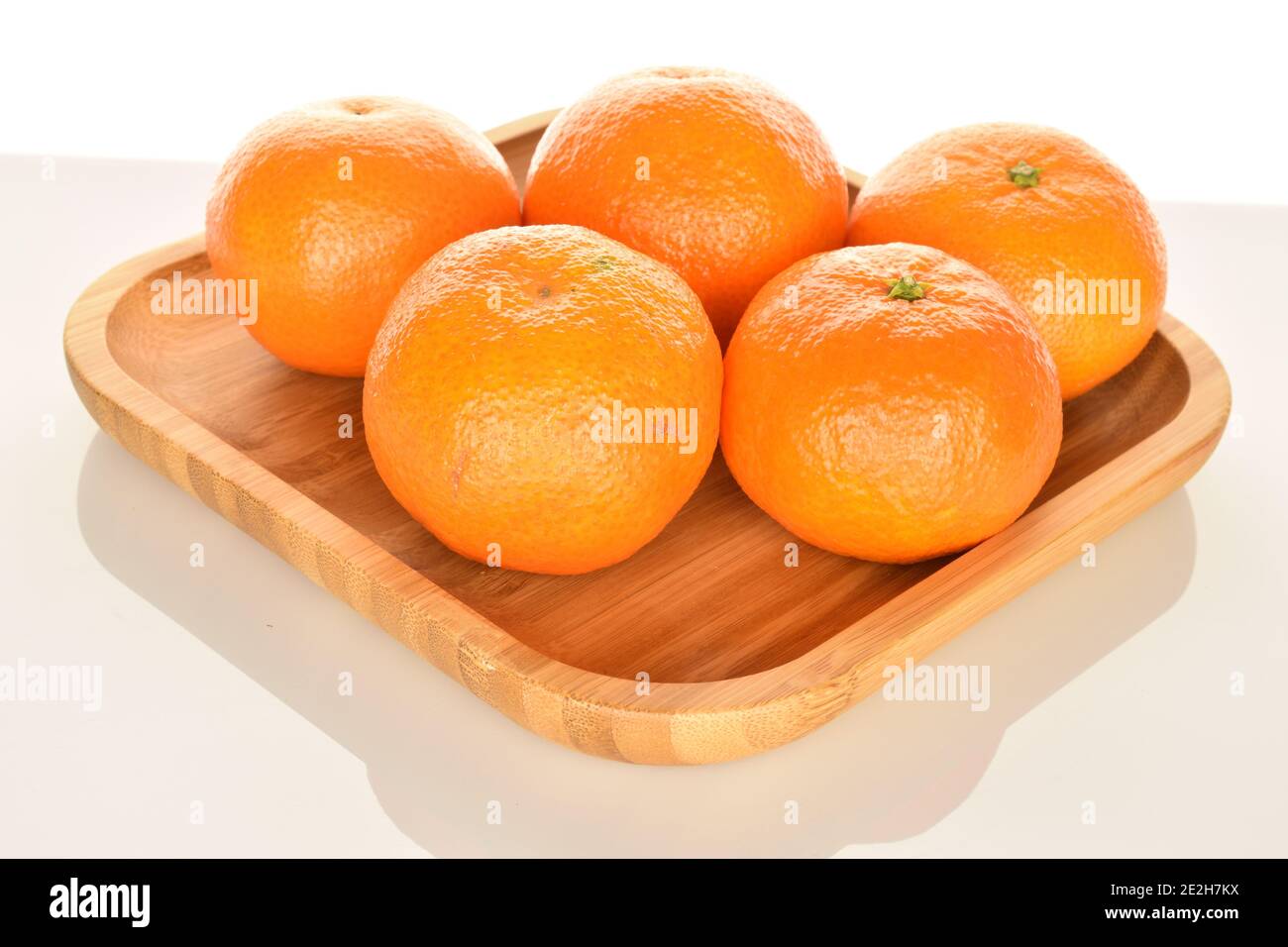 Unas cuantas tangerinas jugosas de color naranja brillante, yacen sobre una  bandeja de bambú, sobre un fondo blanco. La mandarina más cercana, en  primer plano en el foco Fotografía de stock -
