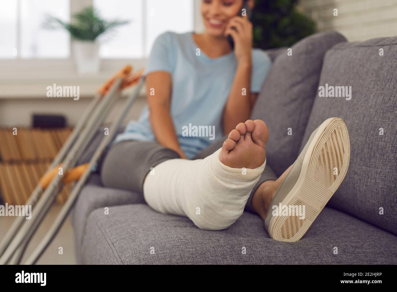 Mujer feliz con una pierna en elenco sentada en el sofá en casa y hablando por teléfono móvil Foto de stock