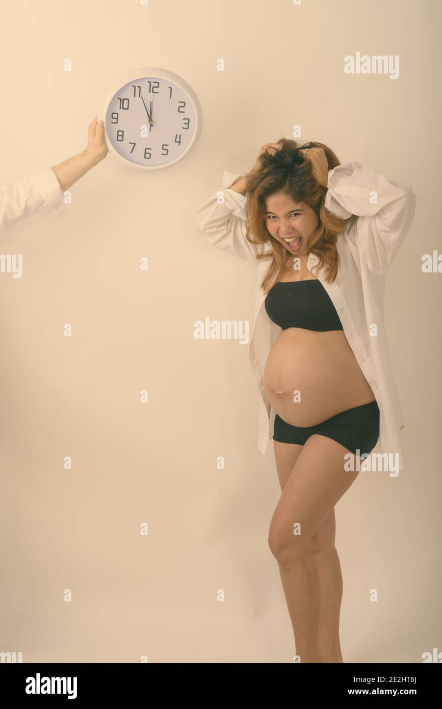 Foto de estudio de jóvenes asiáticos busca mujer embarazada destacó con cuenta regresiva en reloj de pared contra el fondo blanco. Foto de stock