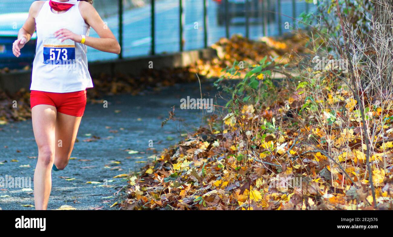 Una niña de secundaria está corriendo en un campo a través carrera en un camino de grava al lado de una carretera bloqueada por una valla con hojas de color otoñal al lado de Foto de stock
