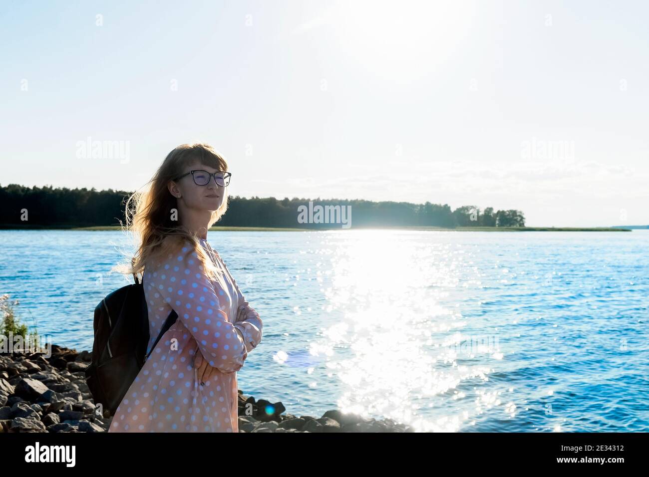 Mujer en vestido ligero se encuentra en una orilla de piedra del río o lago disfrutando de la luz del sol, la relajación y la vista, la salud mental, lejos de todo Foto de stock