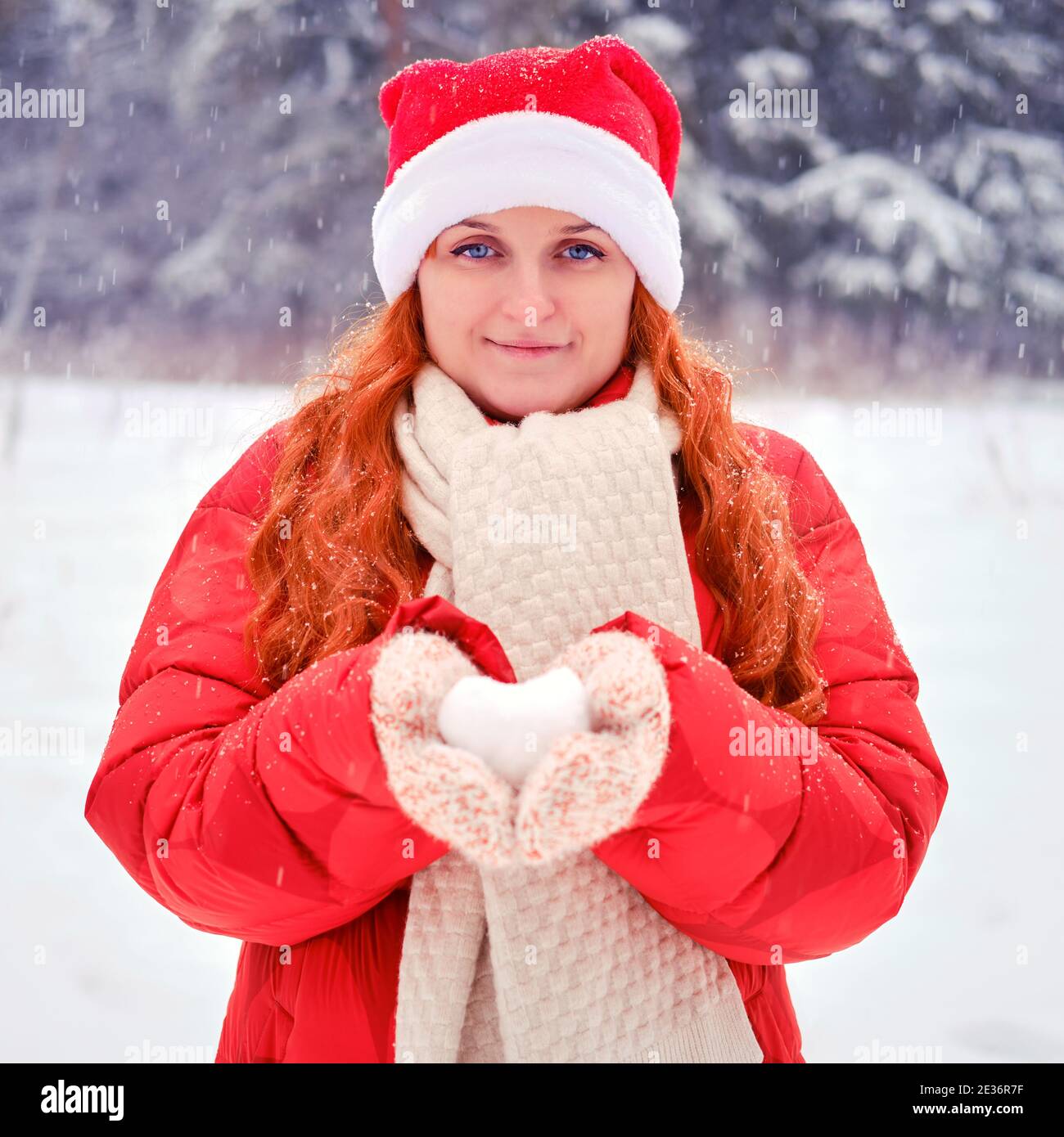 Mujer que sostiene la nieve en forma de corazón en manoplas de punto. Mujer  de pelo rojo en Navidad y año Nuevo camina en invierno con ropa roja  Fotografía de stock -