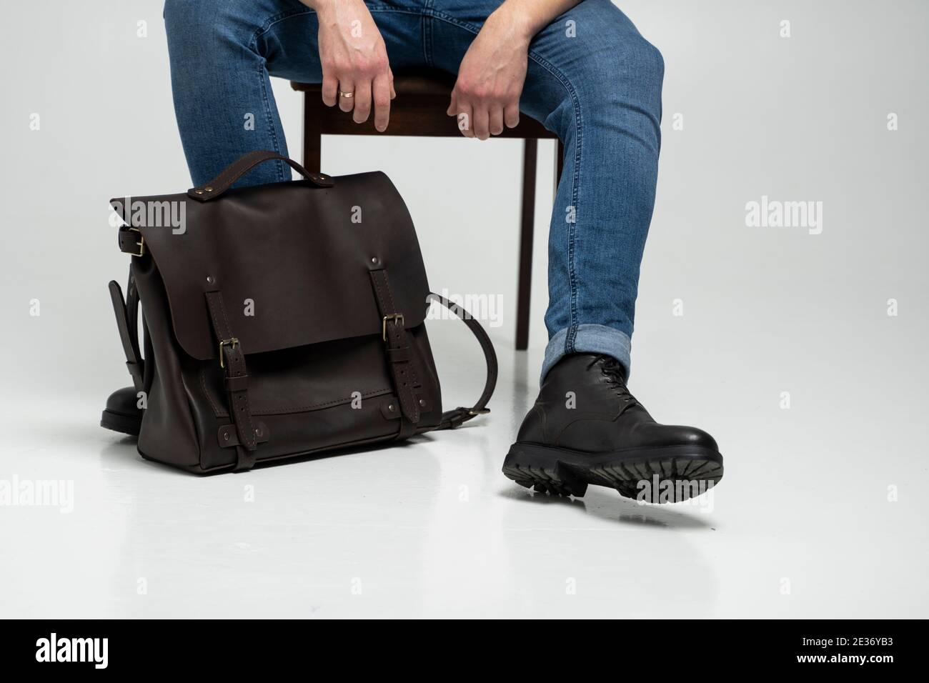 Hombre en un azul jeans y botas negras se sienta en una silla con una bolsa  de cuero marrón hombro para hombre para documentos y portátil en un suelo  blanco. Piel para