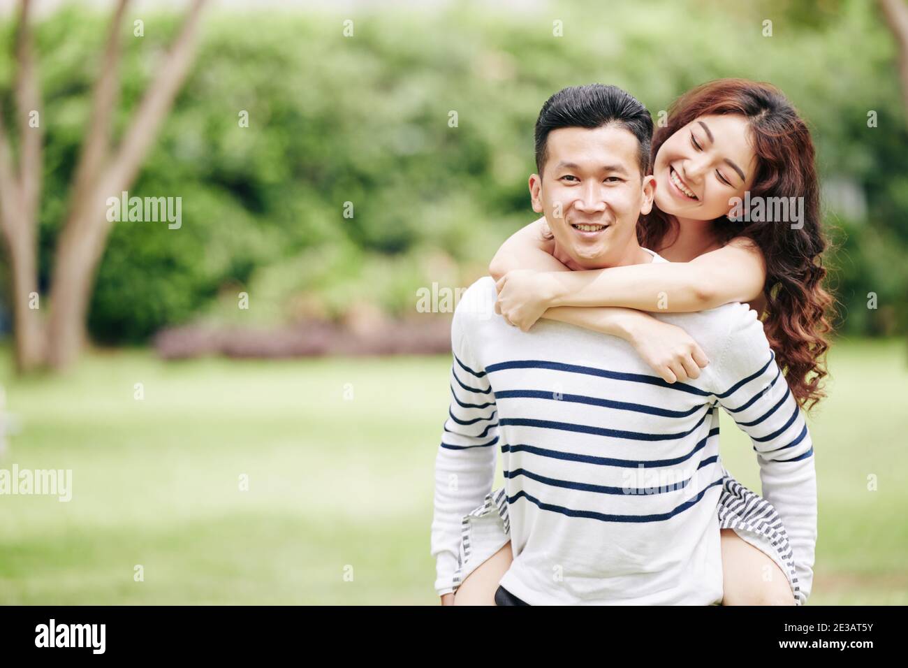 Una joven asiática muy feliz abrazando a su novio desde atrás cuando están de pie en el parque local Foto de stock