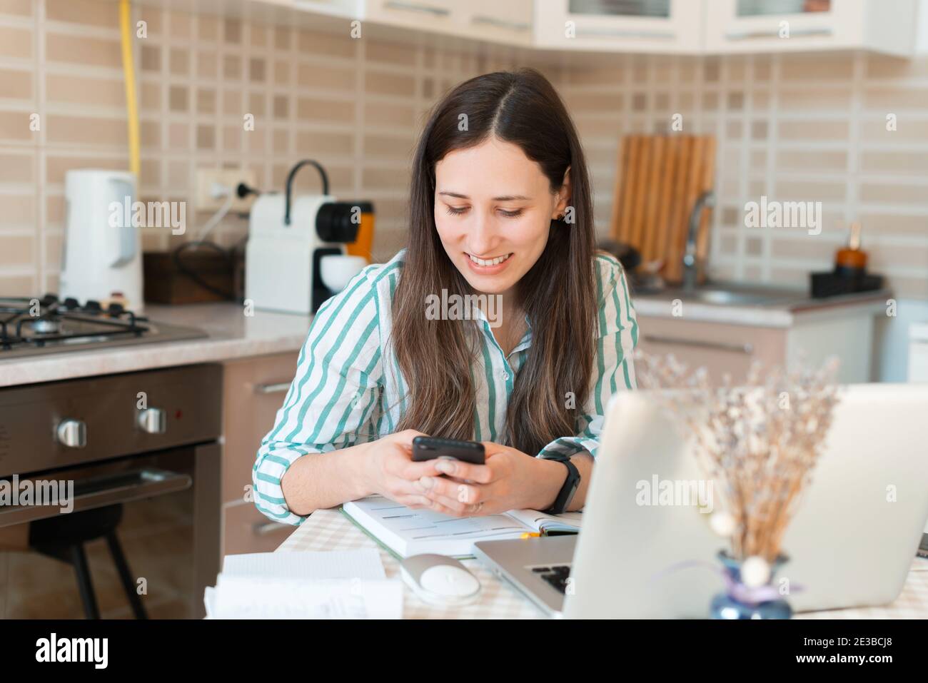 Alegre mujer joven enviando un mensaje a través del smartphone mientras trabaja desde casa en la cocina. Foto de stock