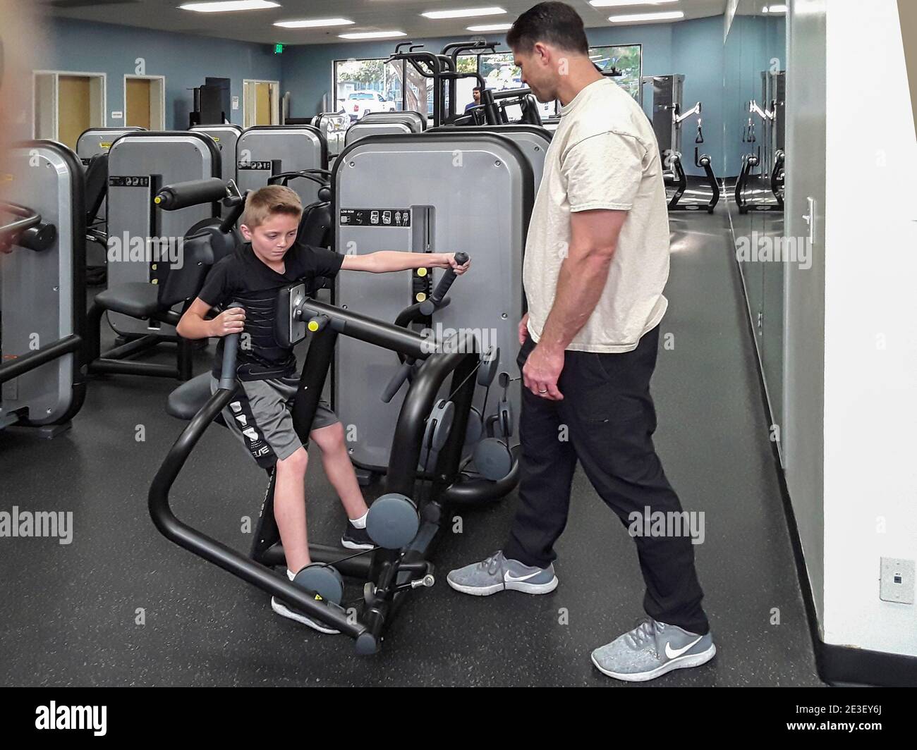 Un padre anima a su hijo mientras trabaja en una máquina de ejercicio en un gimnasio del sur de California. Foto de stock