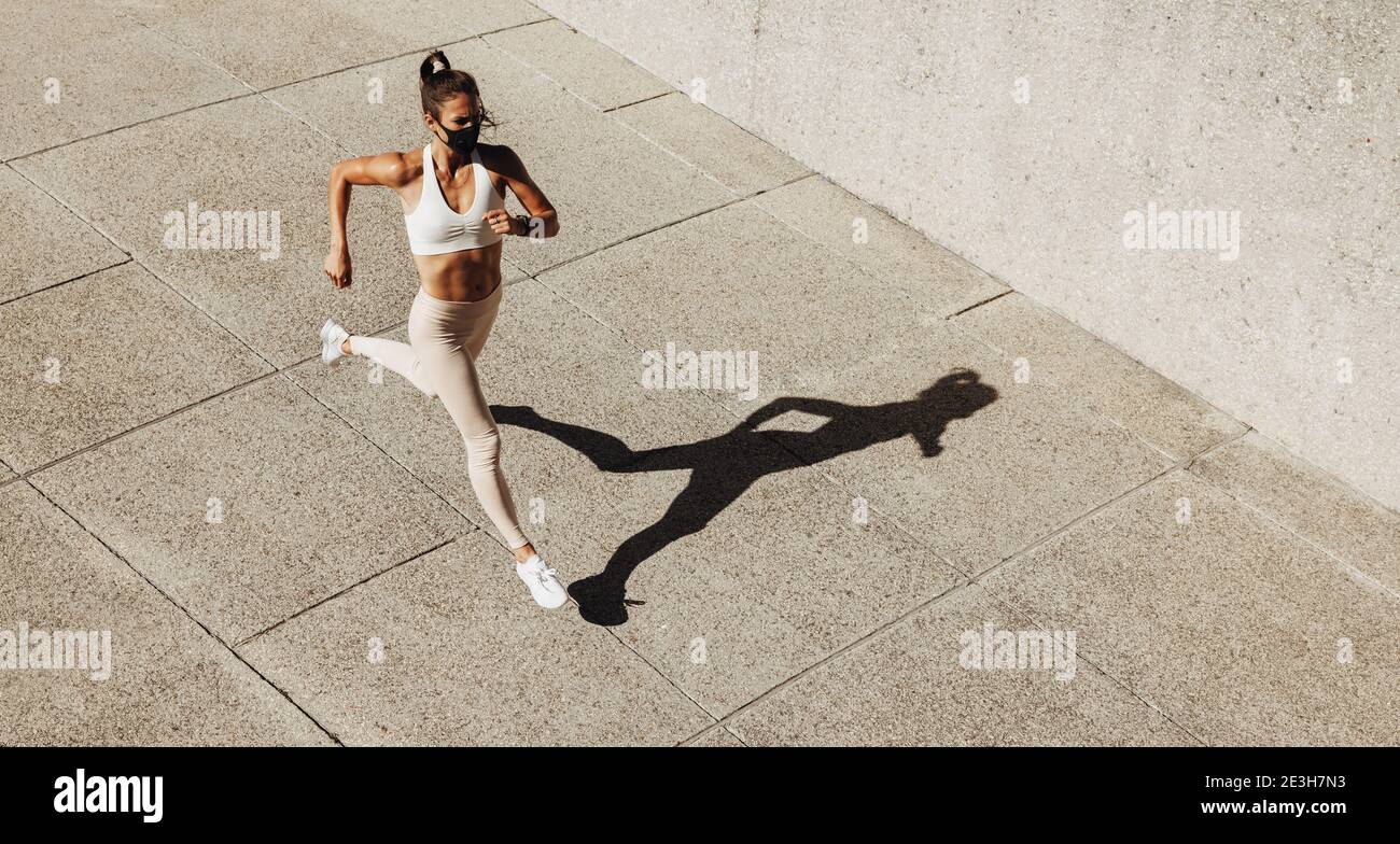Vista en ángulo alto de una mujer que lleva una máscara facial que se enjuaga al aire libre. Mujer corriendo haciendo ejercicio al aire libre usando una máscara protectora. Foto de stock