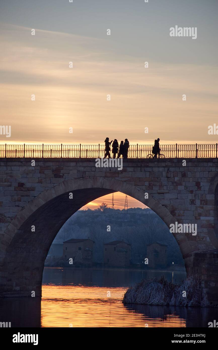 Gente caminando en un puente de piedra al atardecer y los árboles congelados en invierno a la hora dorada. Arcos de piedra, reflejo del sol en el río. Alcantarillas Foto de stock