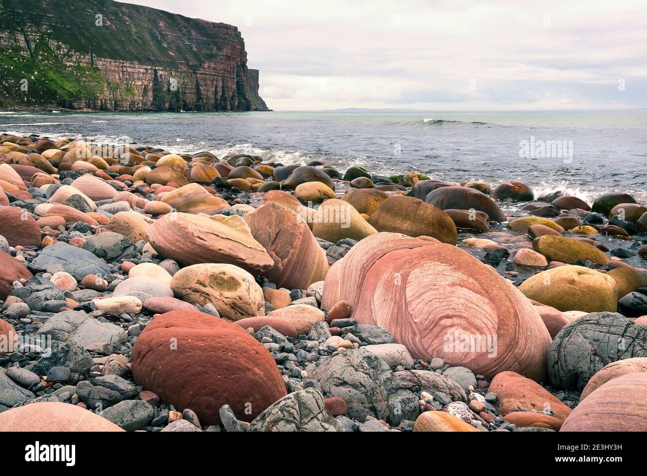 Grandes rocas redondas en la playa escocesa con un acantilado en el fondo En un día nublado en las islas Orkney Foto de stock