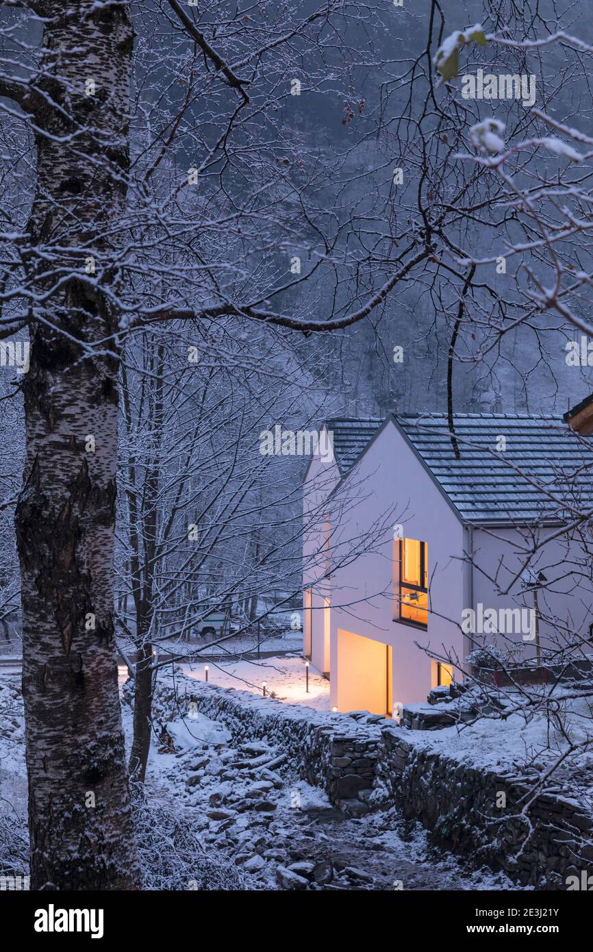 Moderna casa blanca con ventanas luminosas, rodeado de montañas, nieve y árboles. Humor Chirstmas. Nadie dentro. Foto de stock