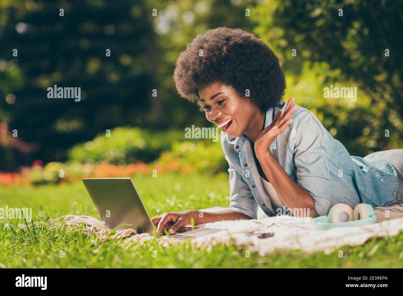 Foto de perfil lateral de la joven estudiante de piel negra acostada en la hierba trabajando con la conversación de la tela de la computadora bromeando riendo Foto de stock
