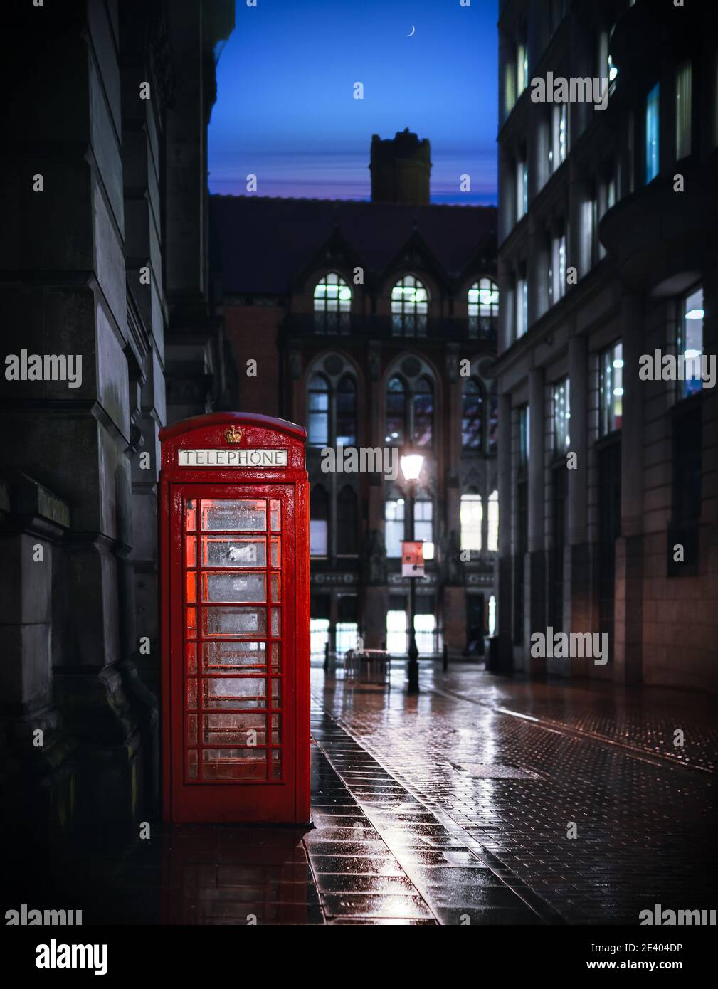 Calle desierta por la noche con el tradicional teléfono británico rojo real la caja iluminó la luz que se refleja en el agua después de la lluvia transparente noche cielo luna vista inquietante Foto de stock