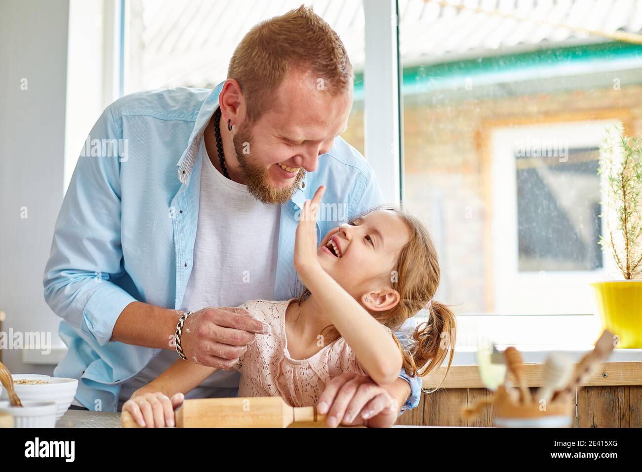 Feliz familia, padre e hija poner masa de galleta en una cocina en casa,  niña se está divirtiendo poniendo un poco de masa en la nariz de su padre  Fotografía de stock -