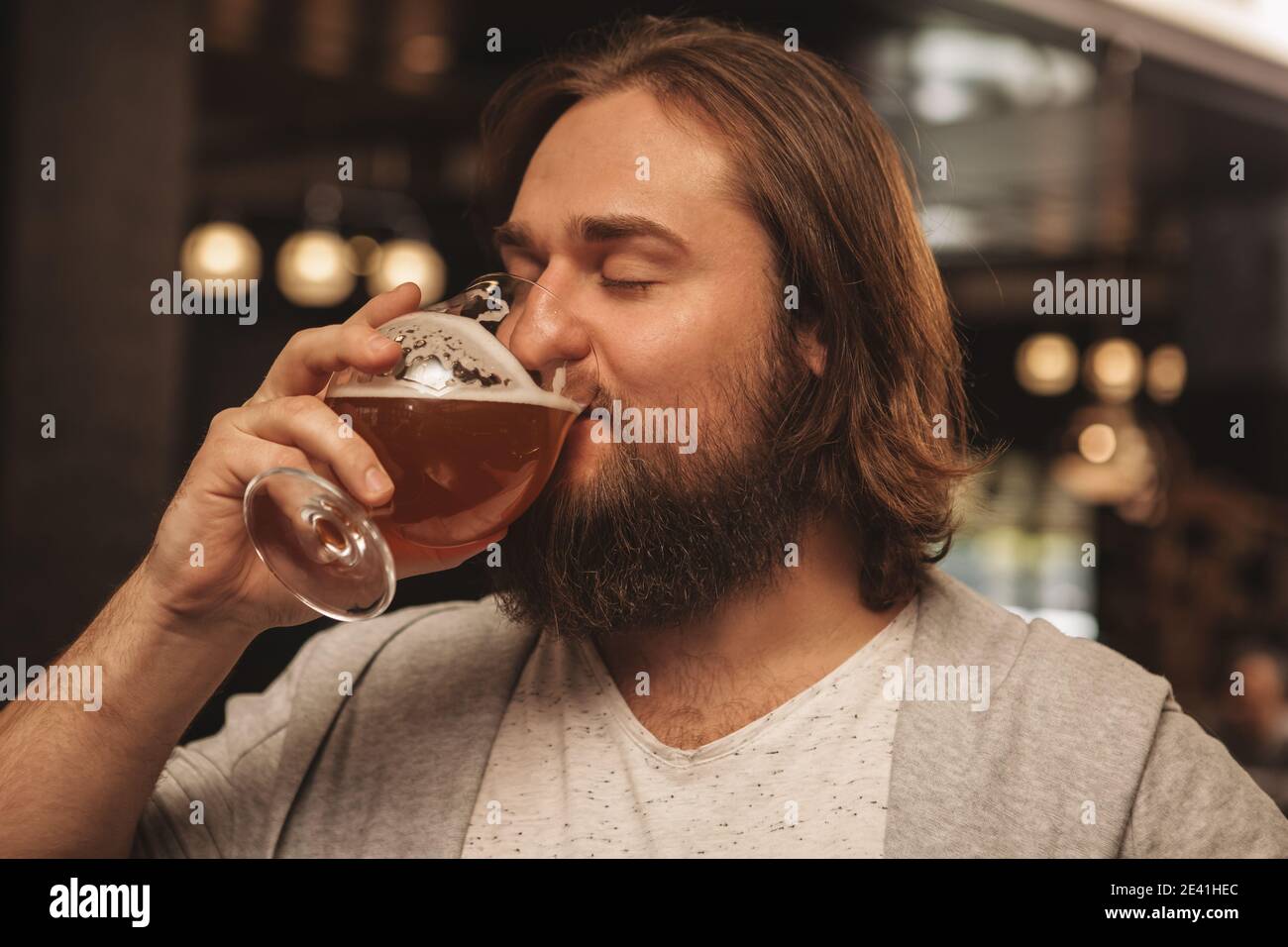 Cerca de un joven barbudo de pelo largo bearded hombre bebiendo cerveza con alegría con sus ojos cerrados en placer. Feliz hombre disfrutando de la deliciosa cerveza fresca en l Foto de stock