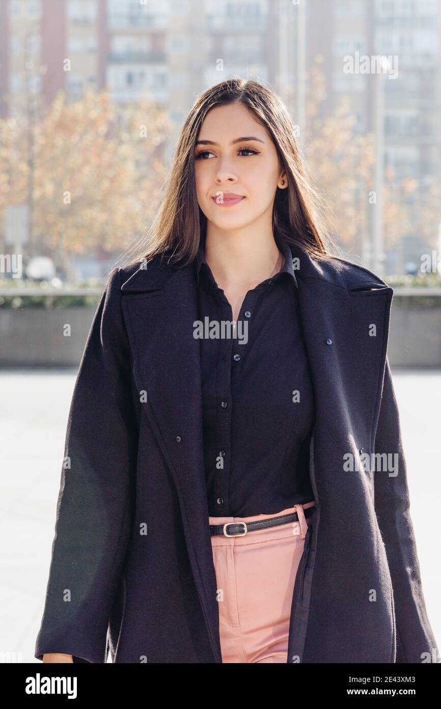 Elegante joven de pelo largo mujer en elegante abrigo negro y tacones altos  caminando en la plaza pavimentada de la ciudad en soleado día de otoño  Fotografía de stock - Alamy