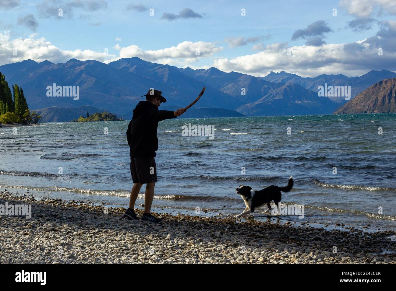 Tiro de un joven con sudaderas con capucha y de pie en la orilla del lago Wanaka con un icónico árbol solitario. Isla Sur de Nueva Zelanda. Foto de stock