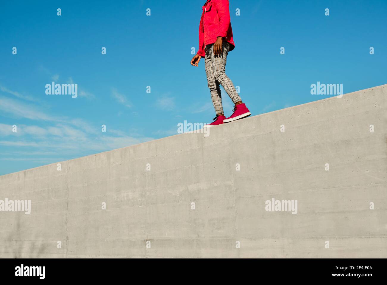 Hombre joven con chaqueta roja y pantalones de cuadros caminando pared de hormigón Foto de stock
