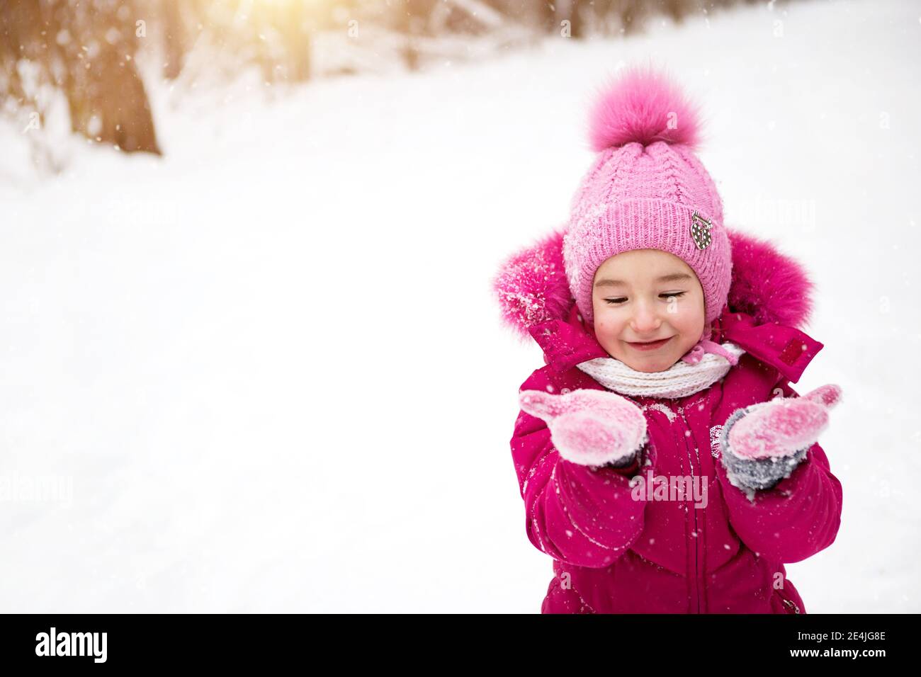 Una niña en el frío mira las mitones de nieve y sonríe. Invierno, caminar  al bebé al aire libre, nieve. Ropa de abrigo, sombrero de punto, pompón de  piel y Fotografía de