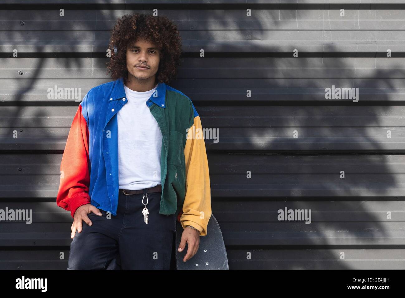 Hombre joven con chaqueta multicolor de pie junto al monopatín pared negra  Fotografía de stock - Alamy