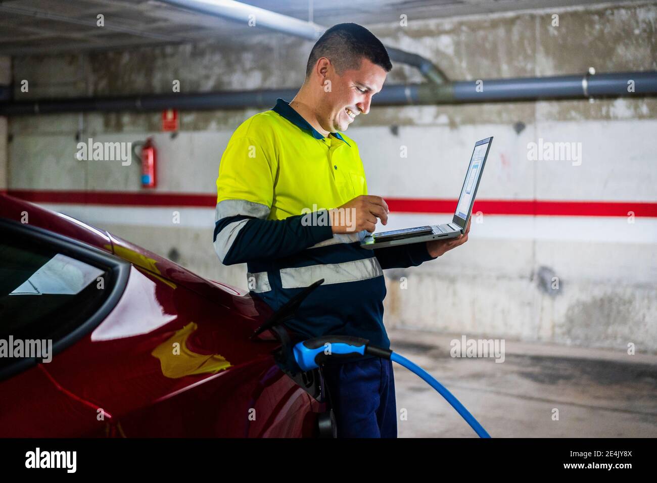 Sonriente programador masculino utilizando teléfono móvil mientras se apoya en eléctrico coche en taller de reparación de automóviles Foto de stock