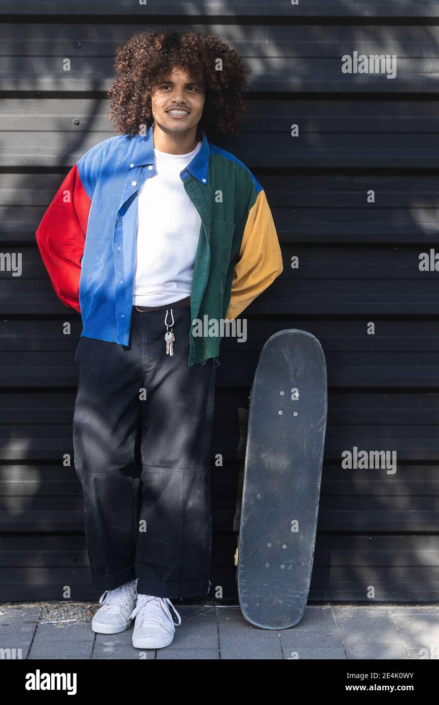 Hombre joven con chaqueta multicolor de pie con monopatín contra pared negra stock - Alamy